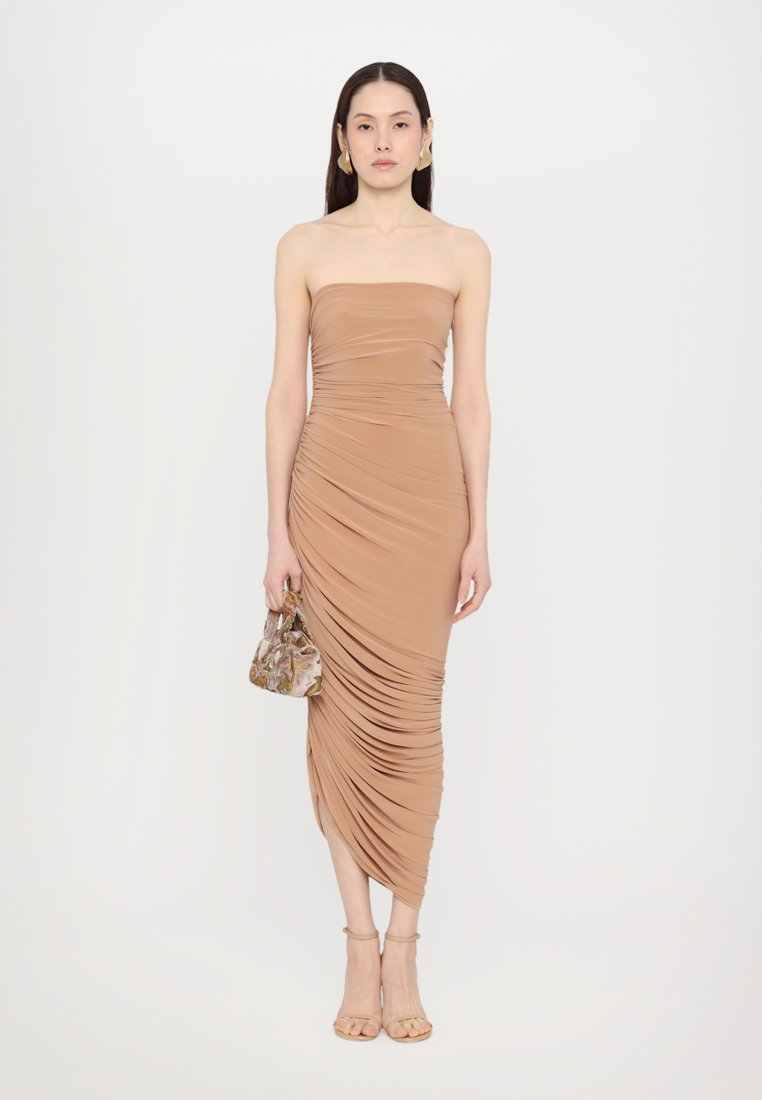 Robe sans manches, longueur genou en tissu beige avec des fronces sur le côté. Présente une coupe ajustée et une texture lisse. Associée à un sac à main à motifs.