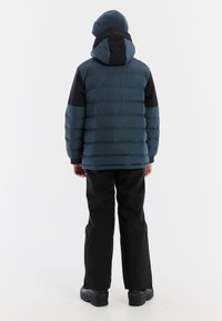 Blau und schwarze Daunenjacke mit Kapuze, mit gesteppter Textur. Getragen mit schwarzen Hosen und stabilen schwarzen Stiefeln, von hinten gesehen.