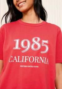 T-shirt rouge en coton avec un col rond, des manches courtes, et un texte imprimé blanc indiquant "1985 CALIFORNIA WESTERN UNITED STATES."
