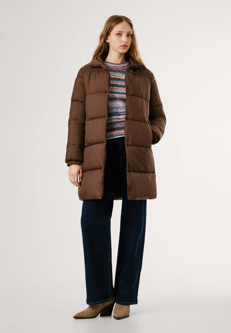 Brązowa kurtka puchowa z kapturem, z pikowanym wzorem, zapinana na guziki z przodu oraz z bocznymi kieszeniami. Nosi ją osoba w paskowanym swetrze i niebieskich dżinsach.