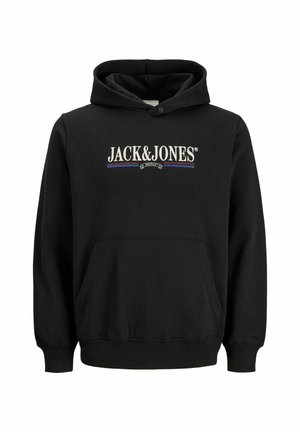 Adolescent aux cheveux bouclés portant un sweat à capuche noir Jack & Jones, un pantalon marron et des chaussures beiges, debout et souriant sur un fond uni.