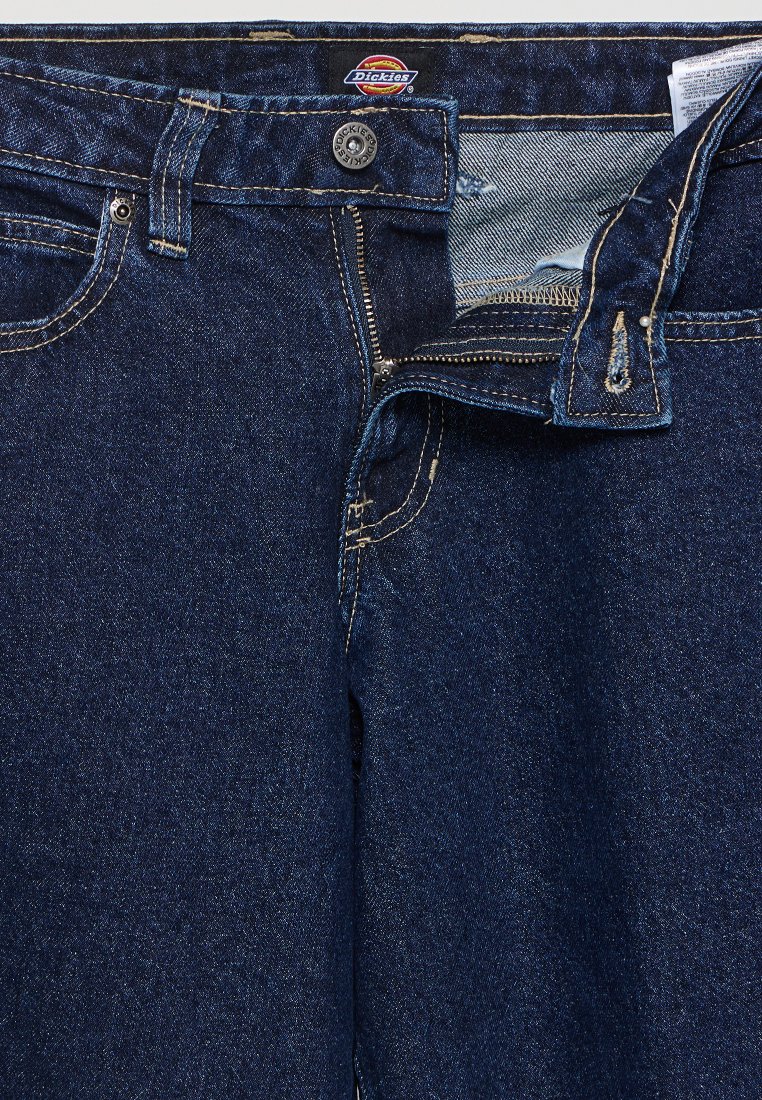 Pantaloni in denim blu scuro con zip, chiusura con bottone superiore e cuciture a contrasto. Etichetta visibile sulla vita, piccoli segni di usura vicino alla tasca.