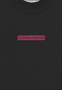 Camiseta de algodón negra con cuello redondo y un logotipo rectangular rosa que dice "CALVIN KLEIN JEANS" en la parte frontal. Textura suave.