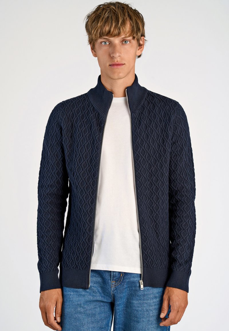 Stickad navyblå tröja med ett strukturerat diamantsmönster och dragkedja fram. Har en hög krage och ribbade ärmslut. Matchas med en vit t-shirt och blå jeans.