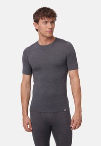 Hombre con cabello rizado corto que lleva una camisa de manga corta ajustada de color gris oscuro y pantalones a juego, mirando hacia su izquierda contra un fondo blanco.