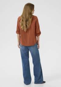Roestkleurig, lang mouwen shirt met opgerolde manchetten, gecombineerd met wijdvallende blauwe jeans. Glad stof; relaxte fit; gebogen onderkant; minimale details.