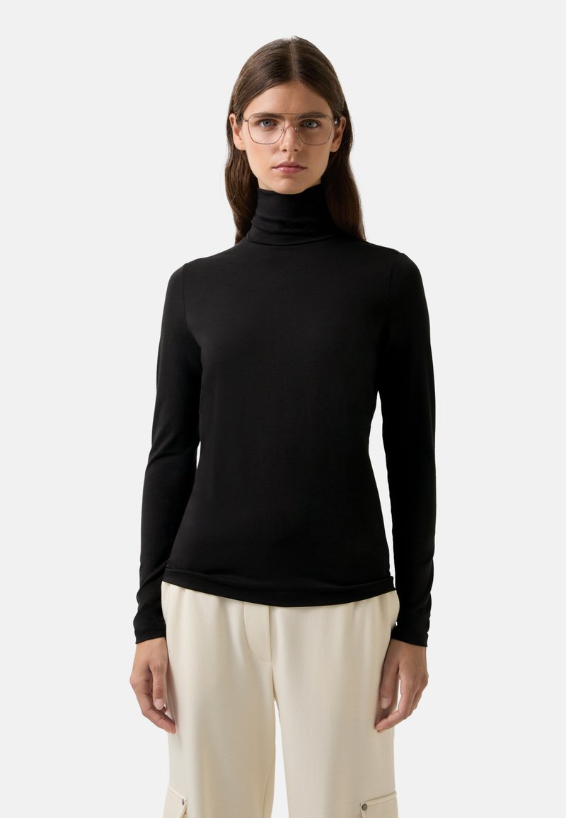 Vrouw met lang bruin haar en duidelijke glazen, gekleed in een zwarte coltrui en beige broek, staande tegen een effen lichte achtergrond.