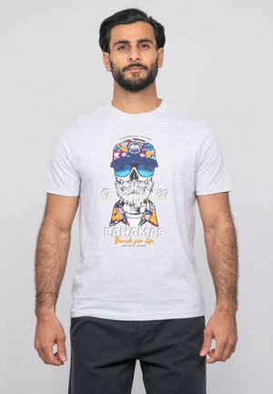 Homme portant un t-shirt blanc avec un graphique coloré de crâne portant des lunettes de soleil et une casquette, debout devant un fond gris clair uni.
