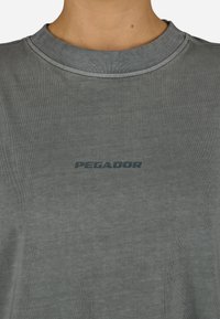 Pelēks ekipāžas kakla T-krekls, kas izgatavots no mīkstas auduma. Tam ir ribota apkakle un neliels, tumšs logs "PEGADOR" priekšā.