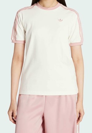 Persoon draagt een wit shirt met korte mouwen met roze bies en strepen op de schouders, gecombineerd met losse lichtroze broek, staand tegen een lichte achtergrond.