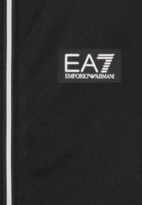 Tissu technique noir mettant en avant un logo EA7 en patch blanc, accompagné de rayures d'accentuation blanches. Texture lisse et design épuré.