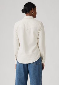 Camisa de manga larga color crema con cuello puntiagudo, dobladillo redondeado, puños abotonados y detalle en la sisa trasera, combinada con pantalones de mezclilla de pierna ancha color azul claro.
