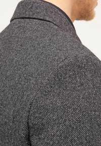 Šedý texturovaný blazer s jemným vzorem herringbone. Disponuje úzkým límcem a štíhlým střihem, který podtrhuje sofistikovaný design.