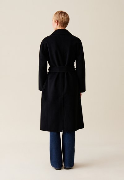 Cappotto di lana nero con vita cinturata, maniche lunghe e colletto a cresta. La parte posteriore presenta uno spacco e una texture liscia. Abbinato a jeans a campana.