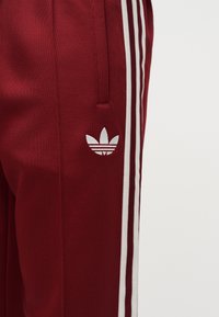 Pantalones deportivos rojo oscuro con el logotipo trefoil de Adidas en blanco y tres franjas blancas que recorren la costura lateral.