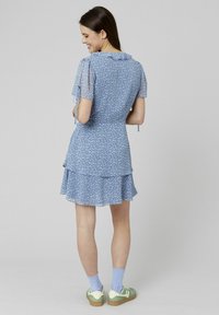 Robe florale bleue avec des manches courtes en tulle, un ourlet à volants et des liens ajustables. Portée avec des baskets vertes et des chaussettes bleu clair.