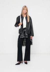 Veste bomber en cuir noir avec une taille cintrée, portée sur un haut blanc, associée à un jean large noir et une chemise à carreaux nouée. Des mules à talons noirs complètent le look.