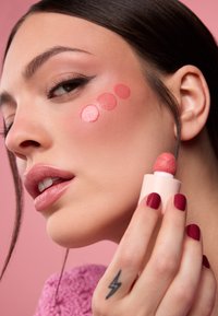 Aplicador de bálsamo para imperfecciones en un envase de color rosa pálido; muestras de rubor coral en la mejilla; piel suave; uñas de color rosa intenso; luces y sombras sutiles.