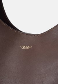 Sac à main en cuir marron avec des surpiqûres visibles autour de la poignée et le logo "COACH NEW YORK" embossé en lettres dorées.