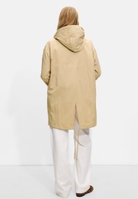 Beige hettejakke med baksliss og justerbar snor. Glatt tekstur; brukt over hvite bukser og fottøy med dyreprint.