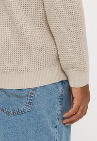 Maglione beige lavorato a maglia con motivo testurizzato, polsini e orlo a coste. Abbinato a jeans di jeans blu con tasca posteriore e dettagli di cuciture.