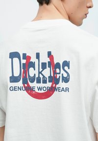 Άσπρο βαμβακερό t-shirt με μεγάλο μπλε και κόκκινο εκτυπωμένο κείμενο στην πλάτη που γράφει "Dickies Genuine Workwear" με γραφικό στοιχείο.