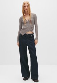 Grå tekstureret indpakningstop med V-hals, parret med mørkeblå vide ben jeans. Bemærkelsesværdig rød syning på jeansene. Flade sko.
