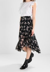 Bílé bezrukávové tričko s volánkovými okraji, kombinované s černou květinovou midi sukni a černými kotníkovými botami. Sukně má splývavý design.