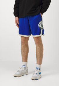 Pantalones cortos de baloncesto azules con ribete blanco, que presentan un gran logo de baloncesto verde. Combinados con zapatillas gris y blancas.