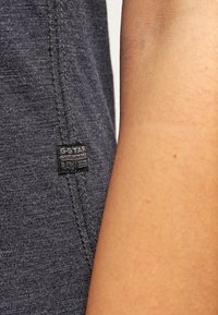 Textil gris de G-Star con un acabado texturizado, presenta una etiqueta negra en la costura lateral, mostrando detalles del logo. Primer plano de la tela y el tono de piel.