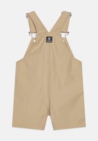 Salopette beige en tissu robuste, dotée de bretelles croisées, de clips en métal ajustables et d'un patch logo sur la poche poitrine.
