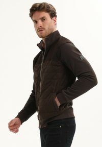 Bruine zip-up jas met een gewatteerde voorkant, gladde mouwen, hoge kraag en zijzakken. Heeft een gestructureerd ontwerp en een logo patch op de mouw.