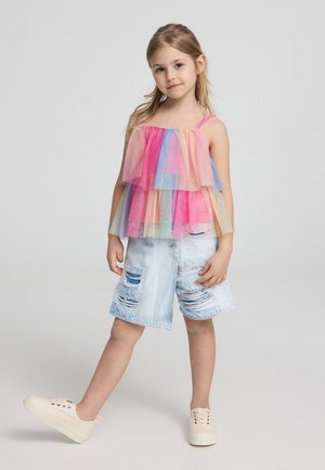 Top senza maniche multicolore con strati di rouches in tulle nei colori rosa, blu e giallo. Pantaloncini di jeans strappati e sneakers beige.