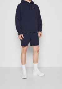 Navy blue hoodie with a front pocket and logo, paired with matching shorts. Worn with white ankle socks and sneakers. Smooth fabric texture.