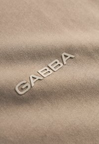 Beiger Stoff mit erhabenen, cremefarbenen Buchstaben, die leicht schräg über das strukturierte Material das Wort "GABBA" bilden.