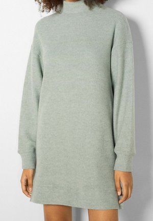 Kvinde iført en langærmet, lysegrå ribstrikket sweater-kjole med høj hals, stående mod en ensfarvet væg.