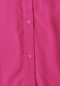 Gros plan sur un tissu de chemise rose vif montrant deux boutons roses cousus et une partie de la patte de boutonnage avec des détails de couture.
