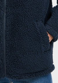 Veste en polaire bleu marine avec une surface texturée, fermeture à glissière et poches latérales. L'ourlet et les poignets présentent un bord doux et arrondi.