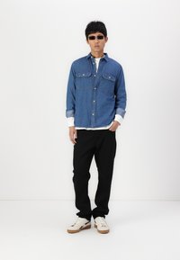 Denim button-up shirt with two front pockets, worn over a white long-sleeve shirt, paired with black pants and cream sneakers.