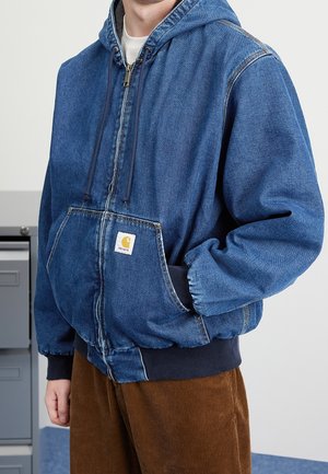 Persoon draagt een blauwe denim zip-up hoodie met een Carhartt-logo en bruine corduroy broek, hand in de hoodie zak.