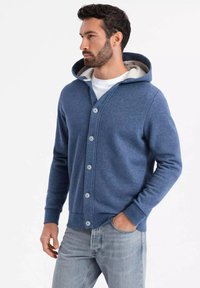 Sudadera azul hecha de tela suave, con un cierre de botones en la parte frontal, forro interior blanco y puños acanalados. Corte casual con un diseño relajado.
