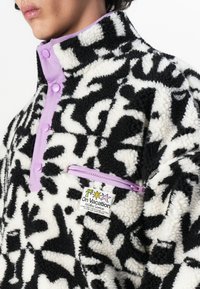 Vlekkige zwart-wit geblokte fleece trui met een lavendelkleurige kraag en zak, voorzien van een label met de tekst "Op Vakantie."