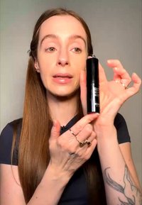 Femme aux longs cheveux tient et montre un produit de maquillage noir KIKO, expliquant ses caractéristiques sur un fond uni.