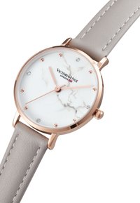 Montre en or rose avec un cadran blanc à motif marbré, un bracelet en cuir gris et des index en cristal. Dispose d'une couronne simple pour les ajustements.