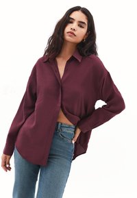 Camicia bordeaux a maniche lunghe con bottoni, dalla vestibilità rilassata, texture morbida e orlo curvo, abbinata a jeans in denim blu.