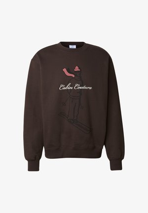 Brun genser med et bilde av en skiløper i rosa og svart med teksten "Cabin Couture." Mykt, teksturert stoff med ribbestrikkede mansjetter.