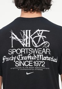 Oseba v črnem T-shirto z belim poškodovanim napisom: "NIKE SPORTSWEAR: skrbno izdelani materiali OD LETA 1972," z logotipom Nike spodaj.