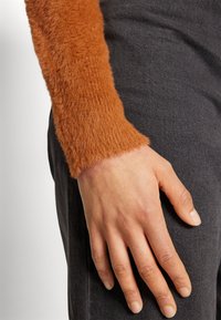 Ręka oparta na ciemnoszarych spodniach, ubrana w pomarańczowy sweter z długim rękawem o miękkiej, puszystej i teksturowanej tkaninie.