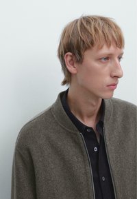 Giacca bomber verde in lana con colletto lavorato a maglia, cerniera argentata e camicia scura sotto, indossata da una persona con capelli corti.