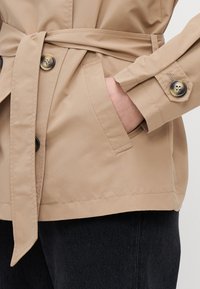 Giacca beige con cintura, realizzata in tessuto liscio, presenta una chiusura a doppio bottone e due tasche laterali. La manica ha un dettaglio con bottone.